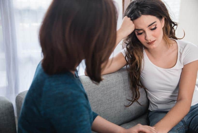 a sad brown haired young woman sitting on a gray couch being comforted by a woman with dark hair wearing a dark green shirt featured image for 85+ Best Resources and Help for Abused Women